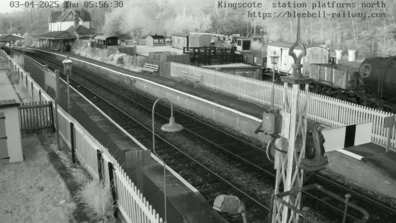 A black and white, high-angle view of a train station platform with railway tracks and several railway cars, under a bright, possibly overcast sky.