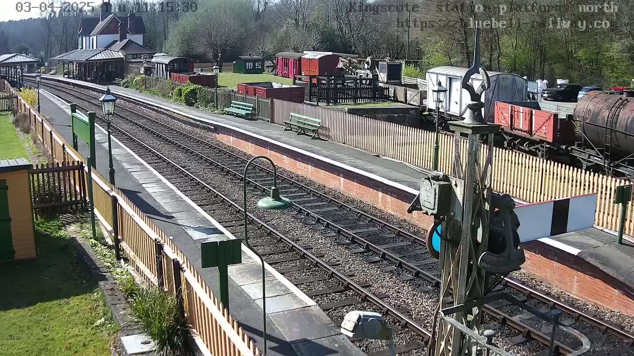 A sunny day at a train station shows railway tracks,  a platform with benches, a station building, and several freight cars.