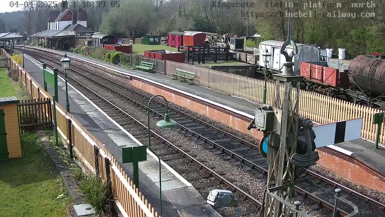 A sunny day at a train station shows multiple railway tracks, a platform with benches, and various railway cars and buildings.