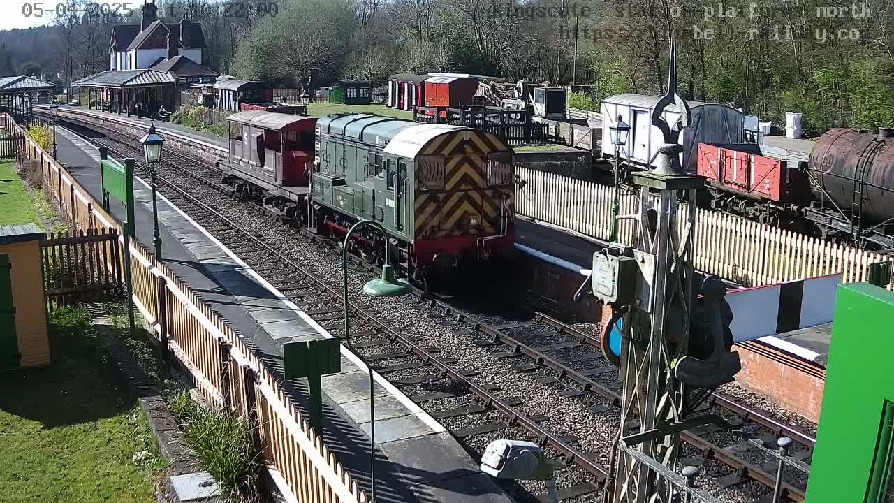 A diesel locomotive leads a train of railway carriages past a station platform on a sunny day.