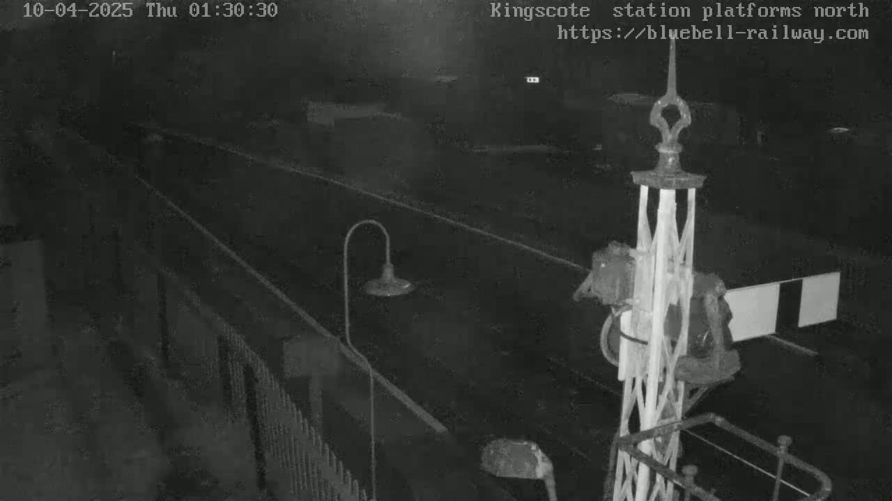 A nighttime, low-light view of a railway platform and tracks, showing a signal and a portion of a fence.