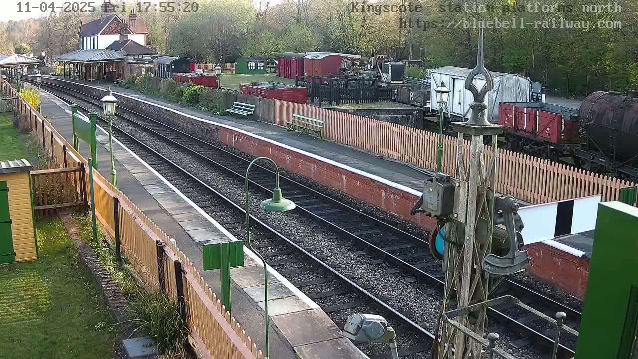 A sunny day at a train station shows two parallel train tracks bordered by wooden fences, with several railway cars and a station building visible.