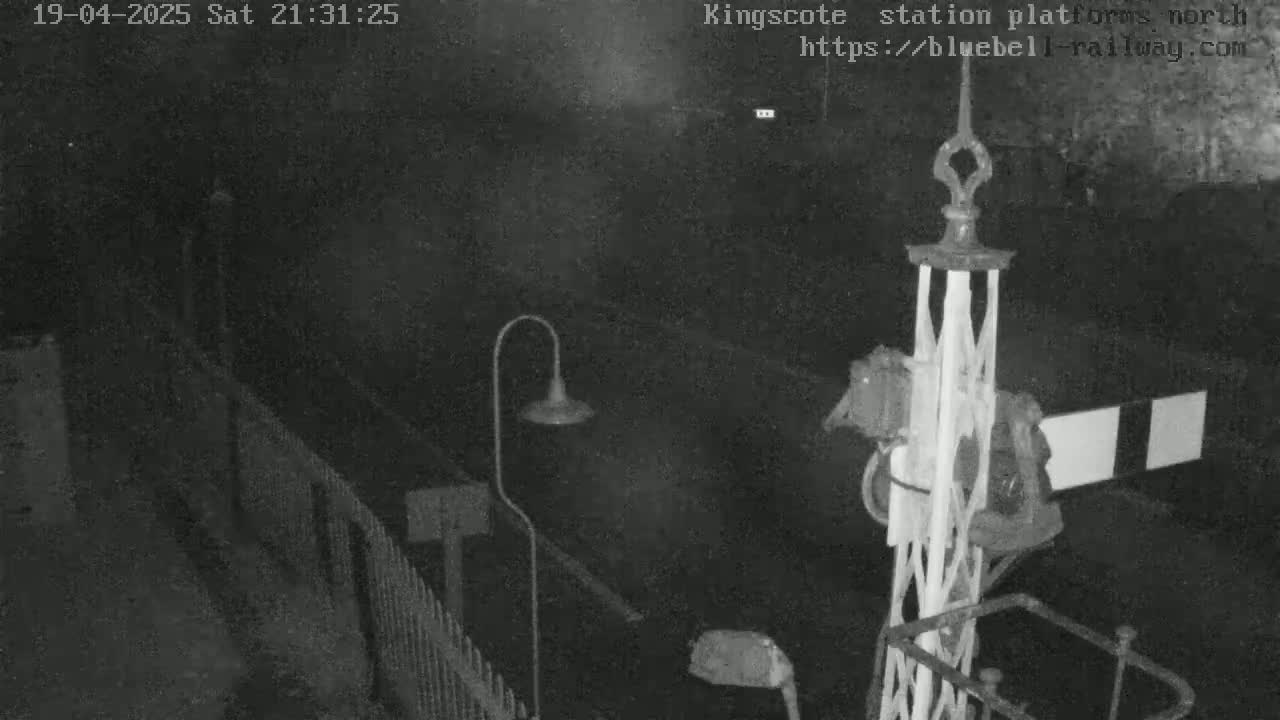 A nighttime, low-light view of a train platform, showing a lamppost, a section of fencing, and a railway signal.