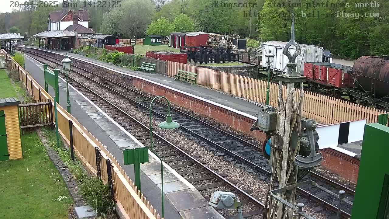 A high-angle view of a train station platform with multiple railway tracks, several railcars, and a station building, all under overcast skies.