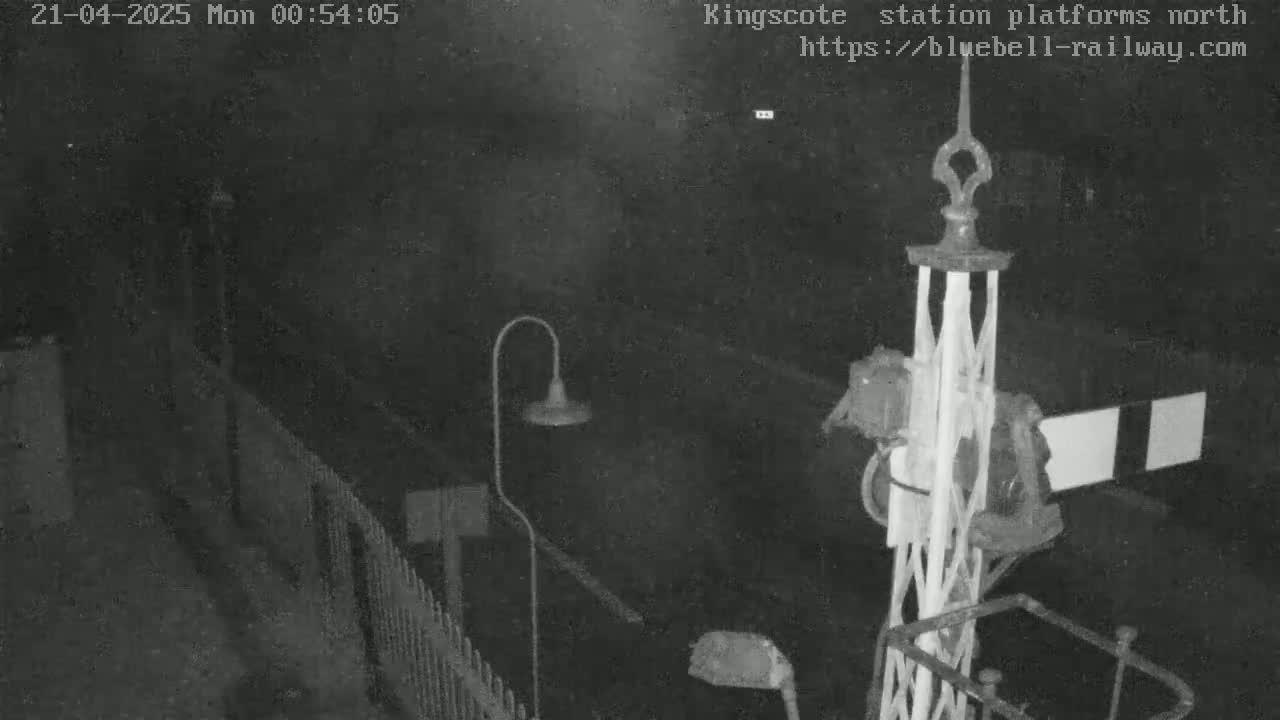 A nighttime, low-light view of a railway station platform, showing a section of fencing, a lamppost, and a railway signal.