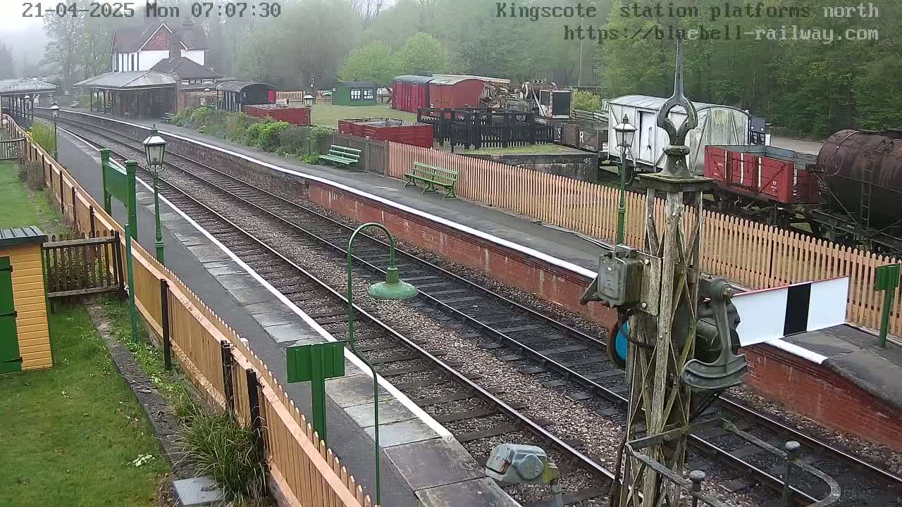 A misty view of a train station platform with multiple railway tracks, several railway cars, and a wooden fence.