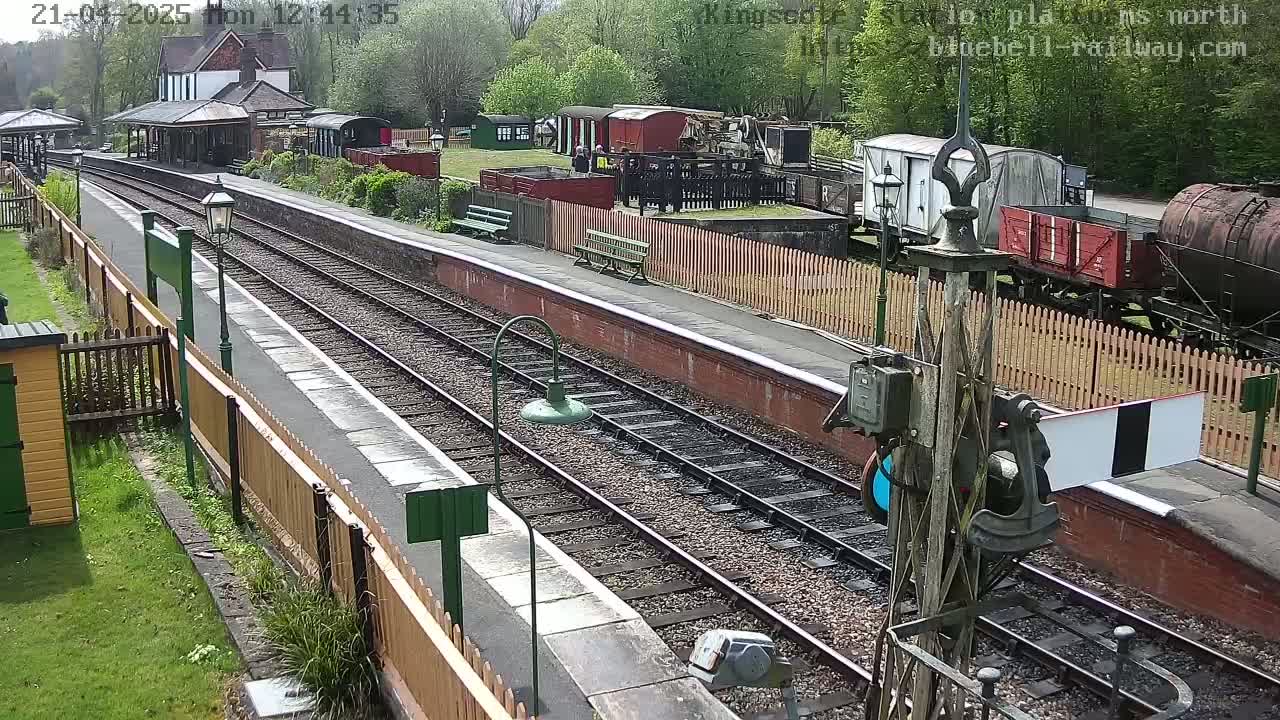 A railway station platform on a sunny day with multiple railway tracks, train cars, and a small station building visible.