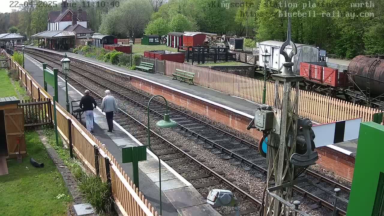 Two people walk along a train platform next to railway tracks and various railway cars and buildings under a partly sunny sky.