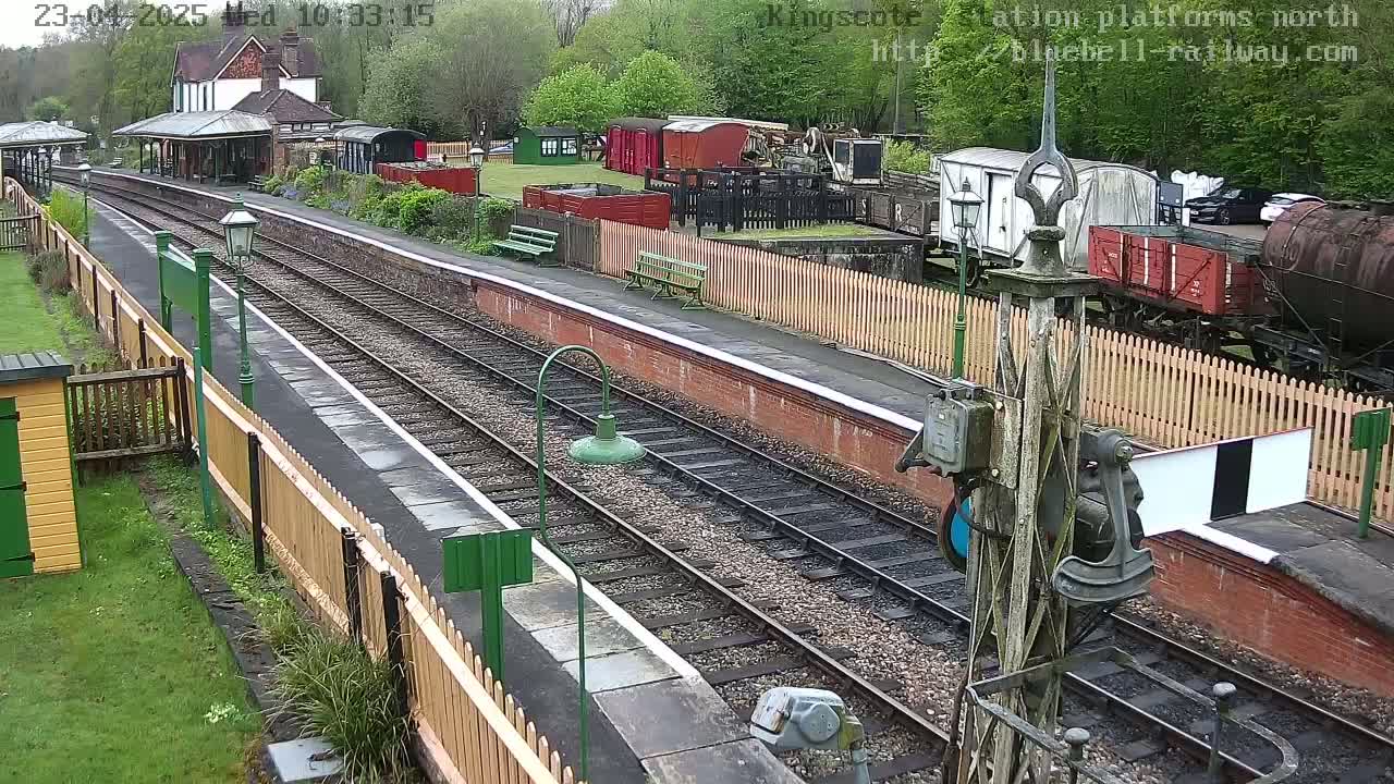 A train station platform with multiple train cars and tracks, surrounded by greenery under a partly cloudy sky.
