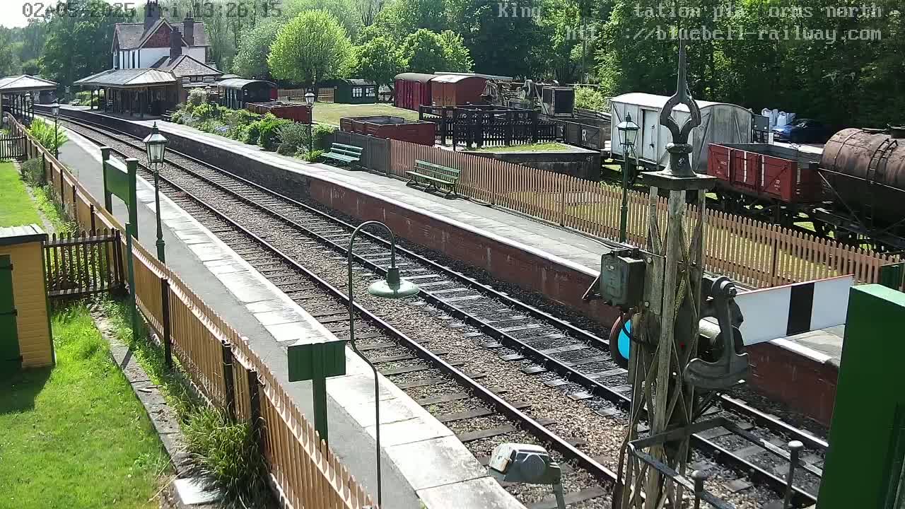 A sunny day at a railway station shows multiple train cars and a station building, with train tracks extending into the distance.
