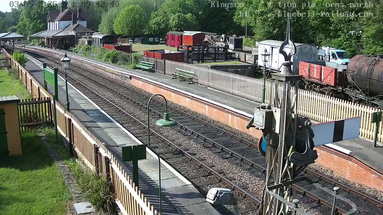 A sunny day at a train station with multiple train cars and a building visible,  railroad tracks running the length of the image, and wooden fences lining the platforms.