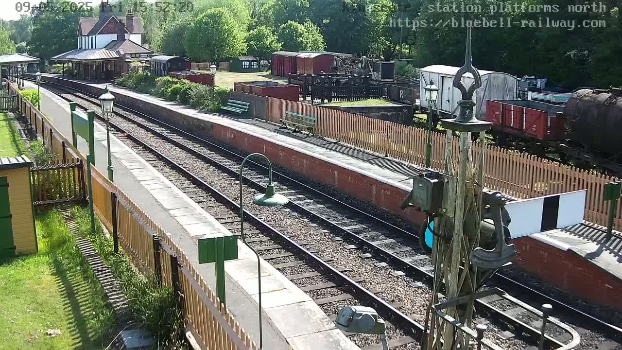 A sunny day at a train station shows two parallel train tracks, a platform with benches and a wooden fence, and several railway cars and buildings in the background.