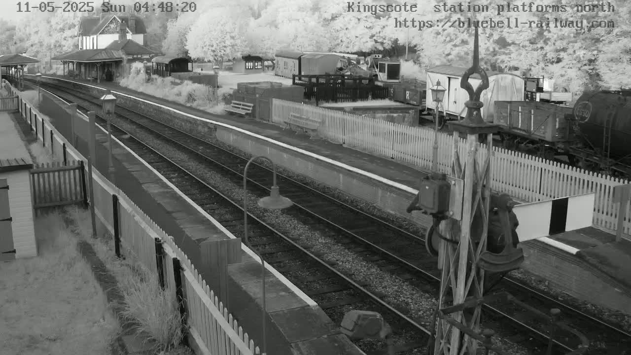 A high-angle, grayscale view of a train station platform with several rail cars and a station building, on a cloudy day.