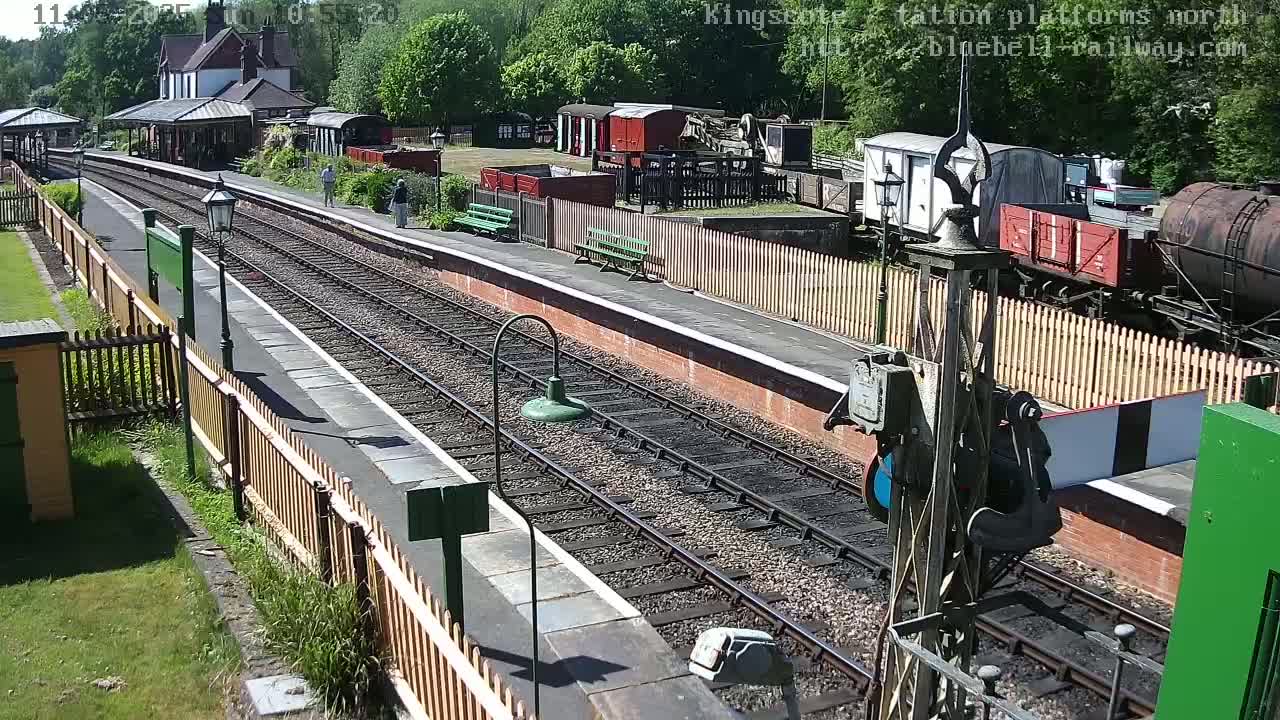 A sunny day at a train station shows railway tracks extending from a platform with several railway cars and a building in the background.