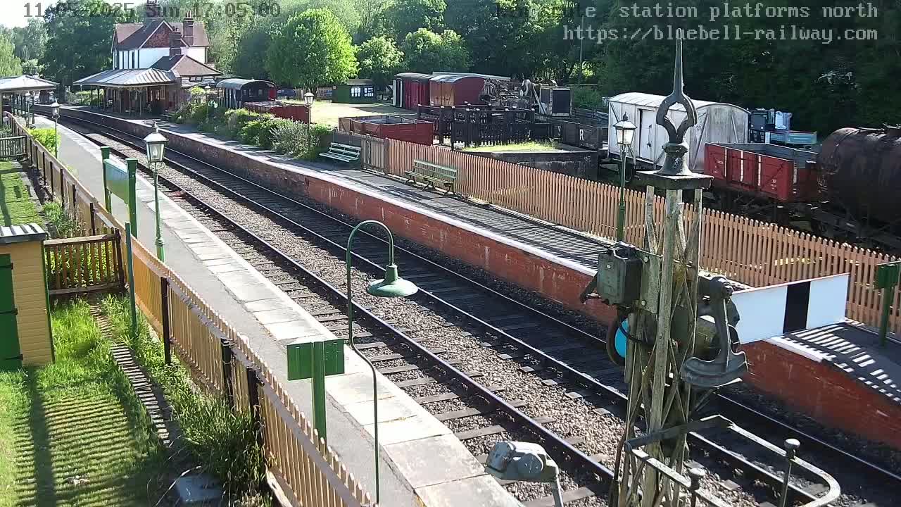A sunny day at a train station, showing railway tracks, platforms, waiting benches, and several railway carriages and buildings.
