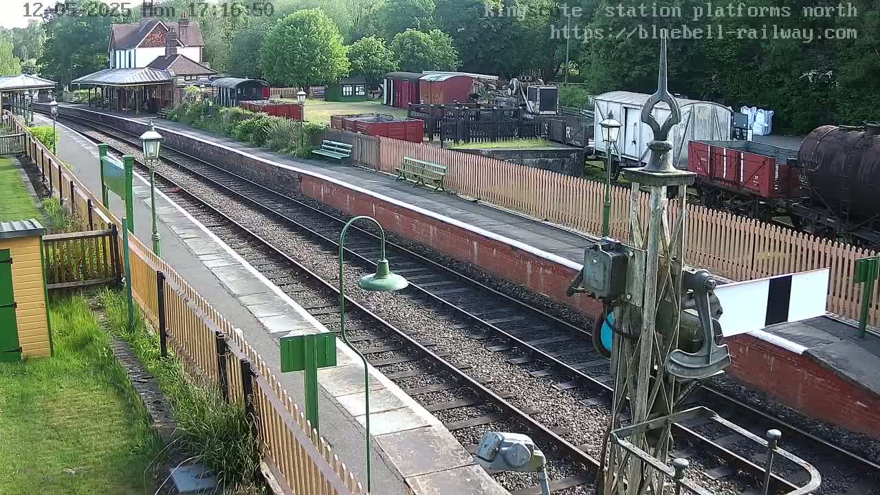 A train station platform with multiple train tracks, railway cars, and a station building is visible on a sunny day.