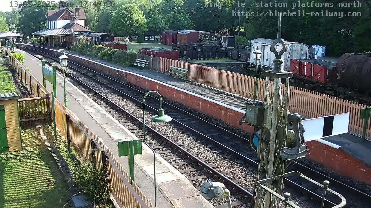 A sunny view of a train station platform with multiple train cars and a station building, surrounded by greenery.