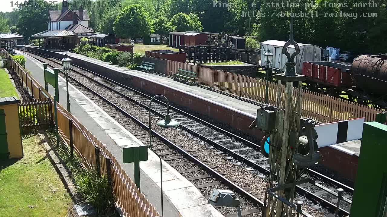 A sunny day at a train station with multiple railway tracks, vintage train cars, and a platform bordered by wooden fences.