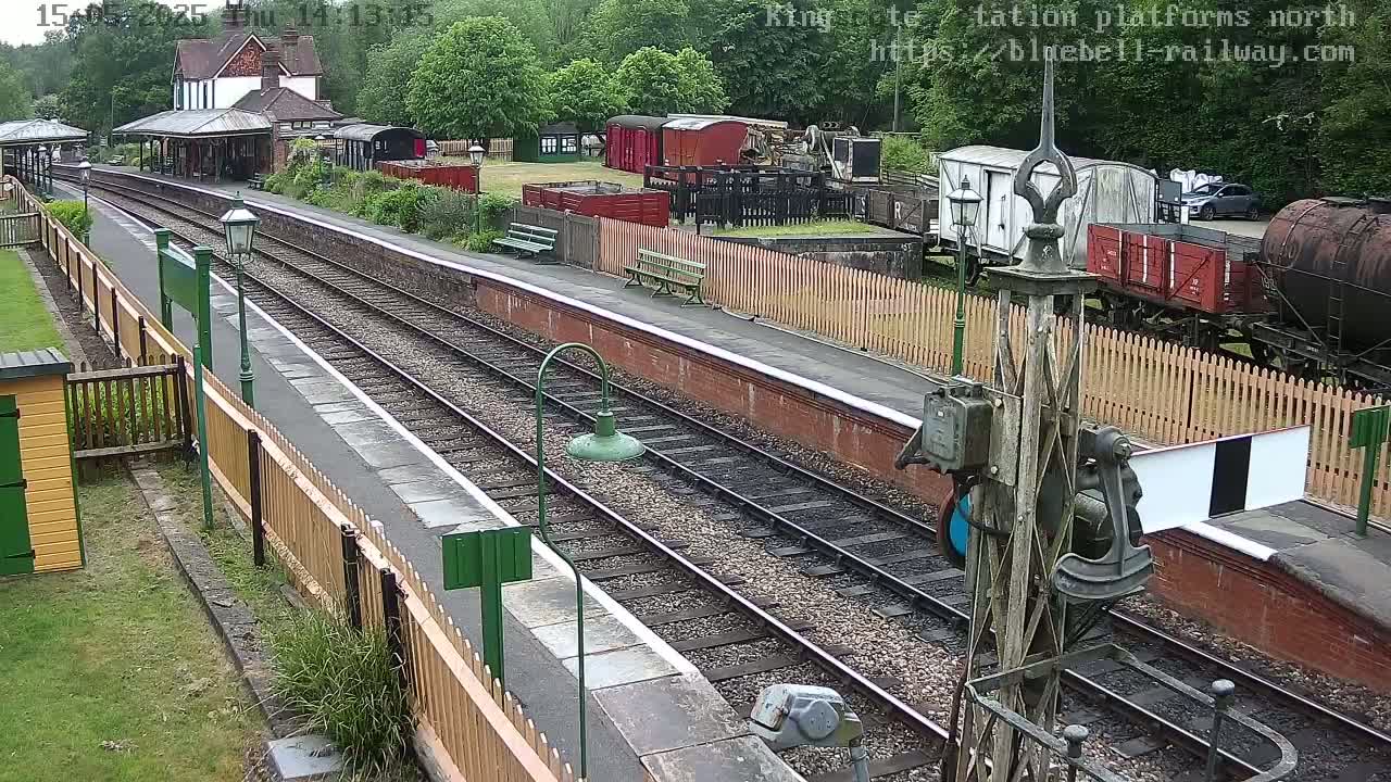 A railway station platform with multiple train tracks, railway cars, and a station building is seen on an overcast day.
