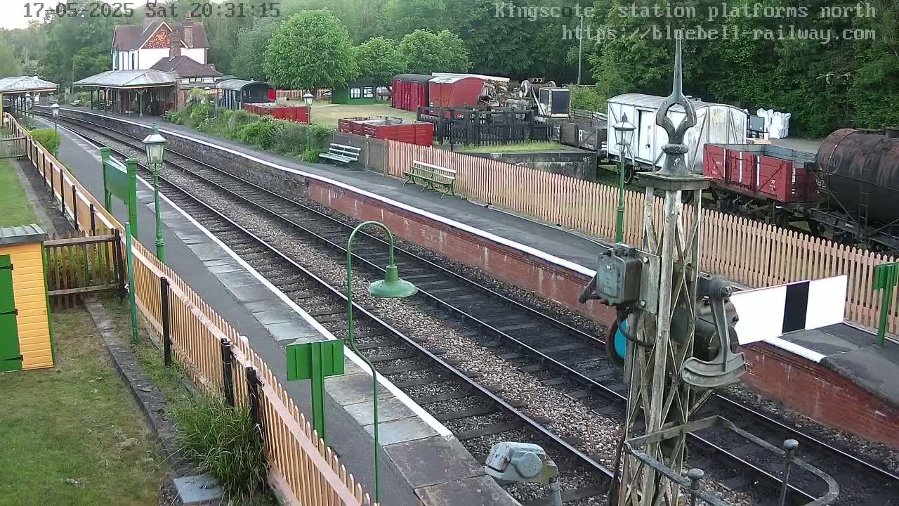 A train station platform with two parallel train tracks, several railway carriages, and a wooden fence separating the platform from the railway yard; the weather appears overcast.