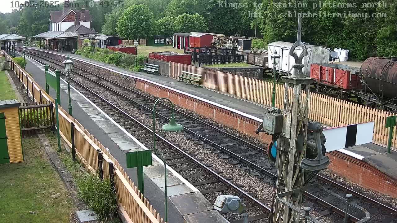 A railway station platform with multiple train cars and tracks visible, on a partly sunny day.