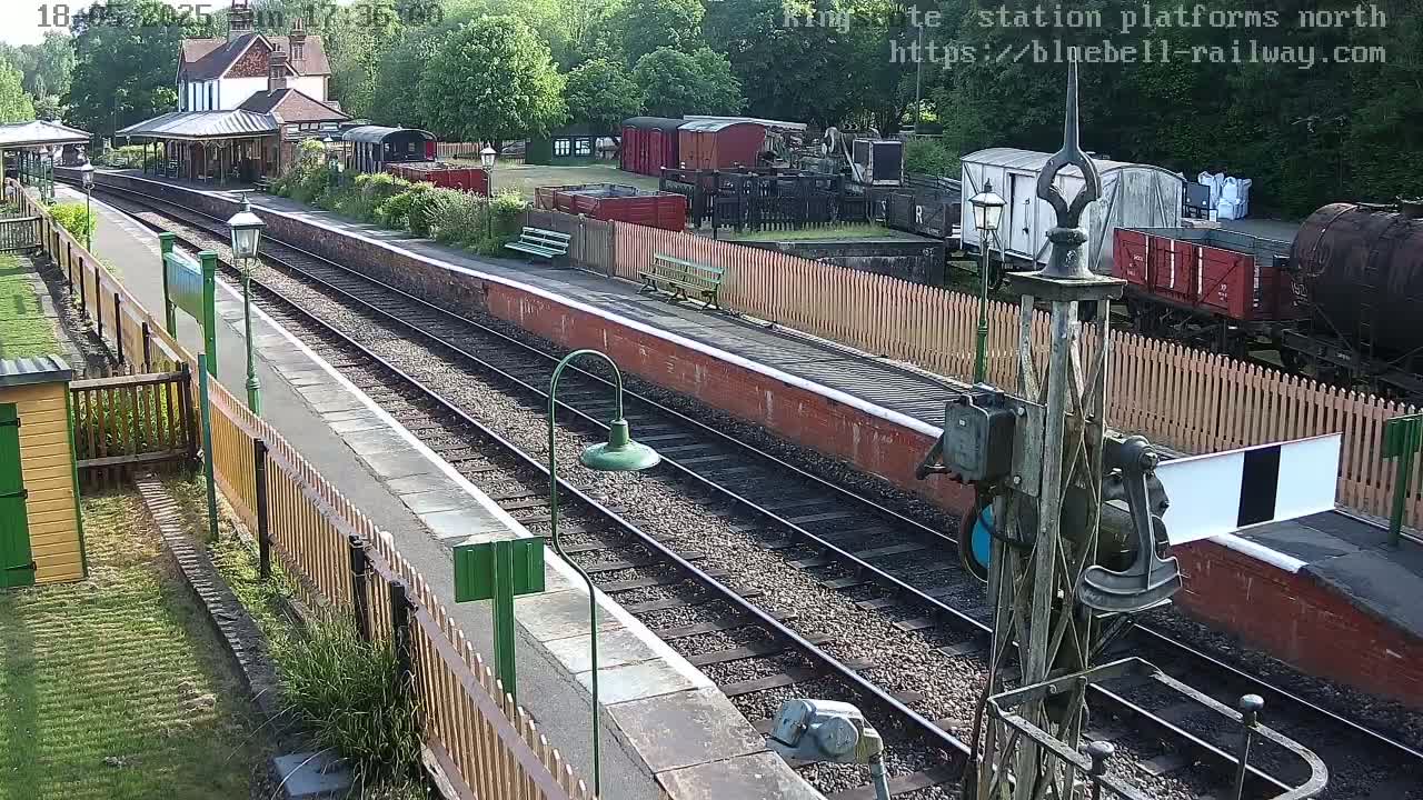 A sunny view of a railway station platform with multiple train cars and tracks, surrounded by wooden fences and greenery.