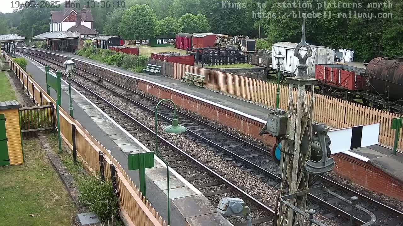 A railway station platform with multiple train cars and tracks, surrounded by wooden fences and greenery, on a partly cloudy day.