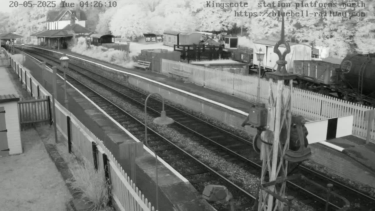 A grayscale overhead view of a train station platform with railway tracks, several train cars, a station building, and fences, on a slightly overcast day.
