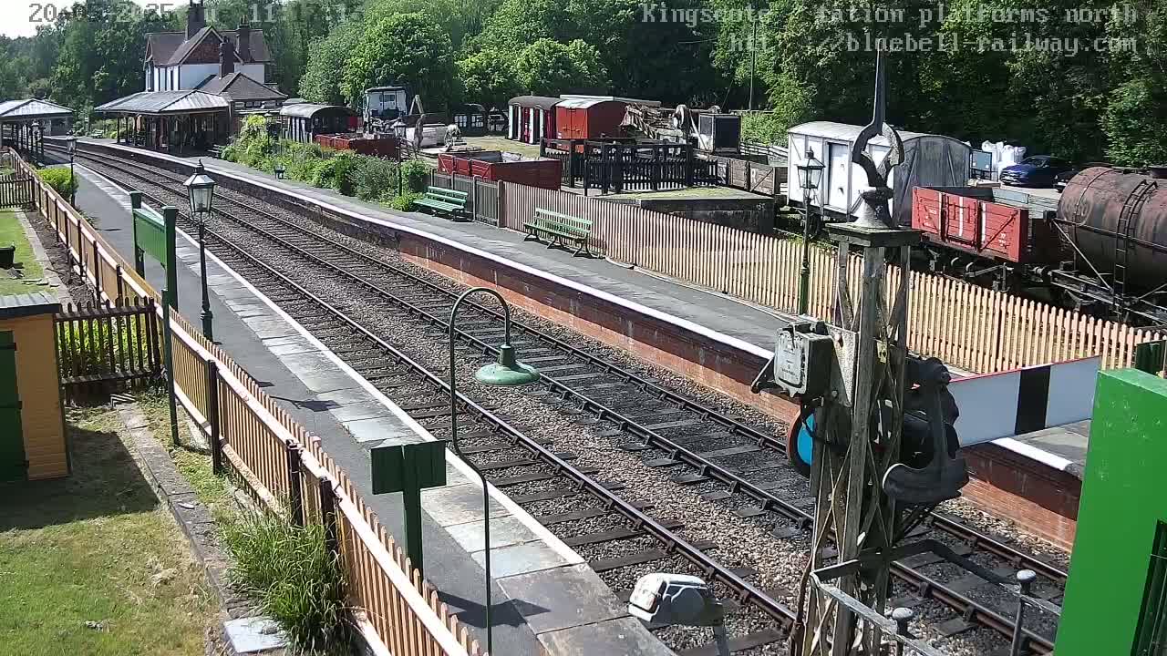 A sunny day at a train station with multiple train cars and rail lines visible, surrounded by a wooden fence and greenery.