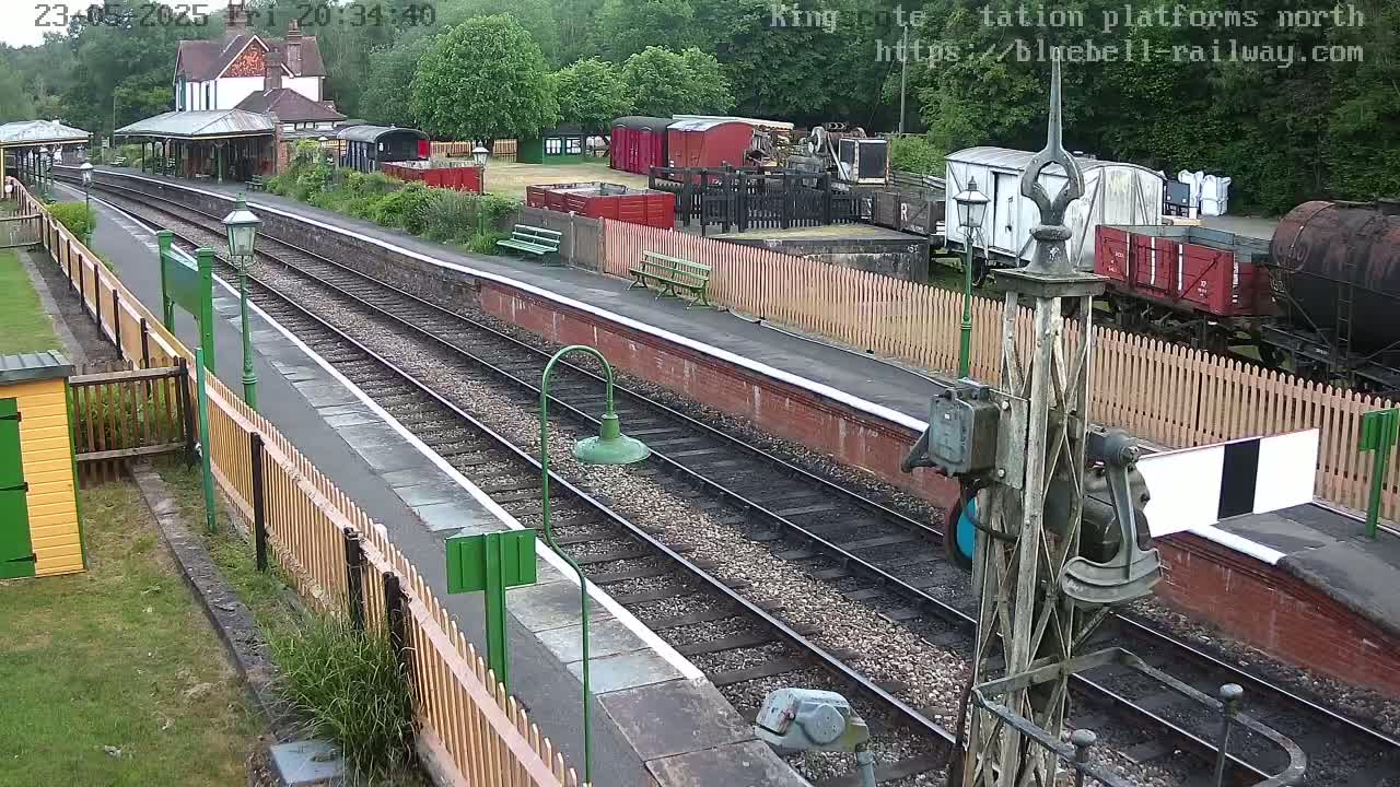 A railway station platform with multiple train tracks, railway cars, and a building in the background on a partly cloudy day.
