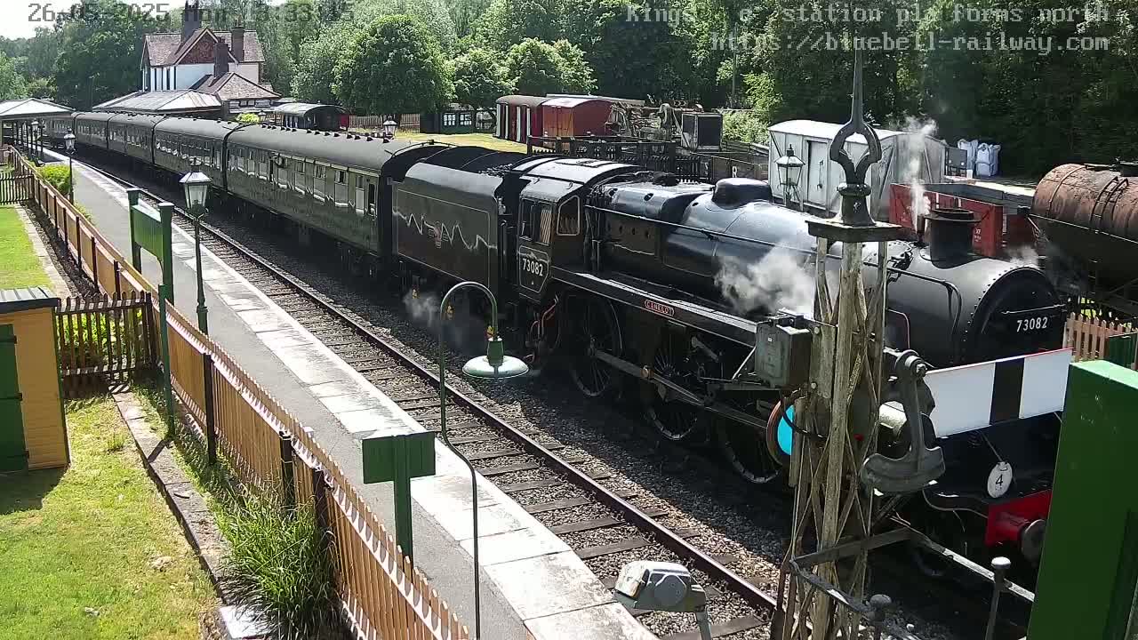 A black steam train pulls into a railway station on a sunny day.