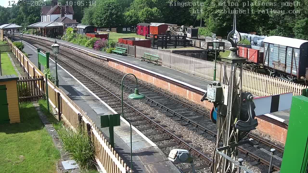 A sunny view of a train station platform with several railway carriages and tracks, and a station building in the background.