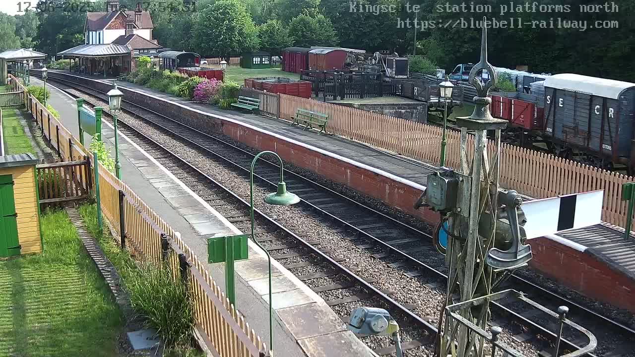 A railway station platform with train tracks, railway cars, and a station building under a sunny sky.
