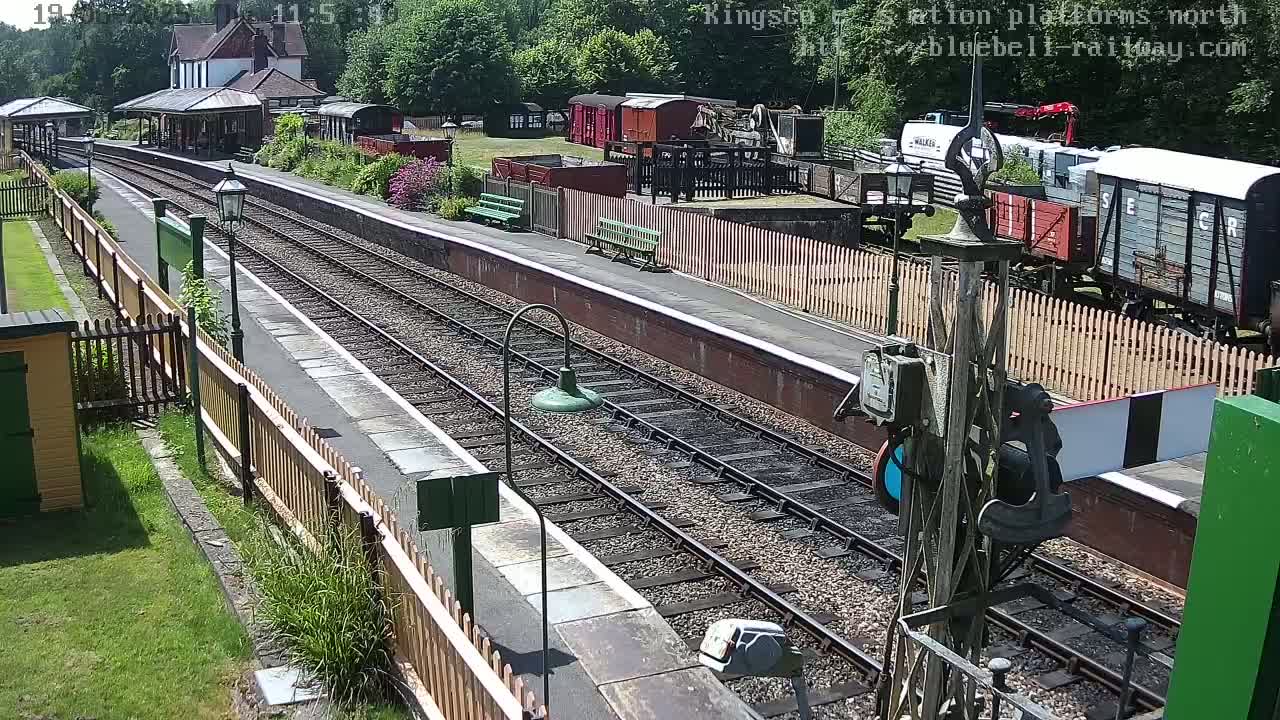 A sunny view of a railway station platform with several train cars visible on the tracks, and a station building in the background.