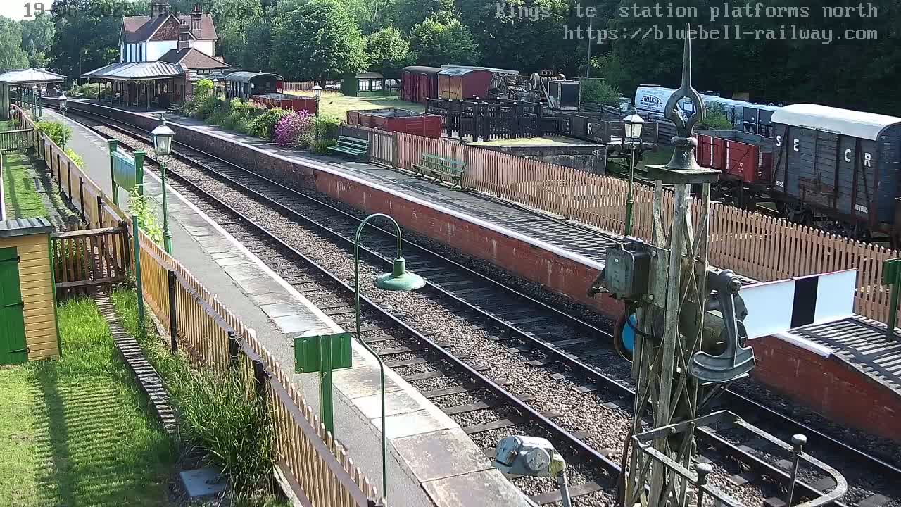 A sunny view of a train station with multiple railway tracks, several freight cars, a station building, and wooden fences.