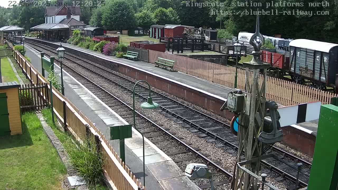 A railway station platform with train tracks, several railway cars, a station building, and wooden fences on a sunny day.