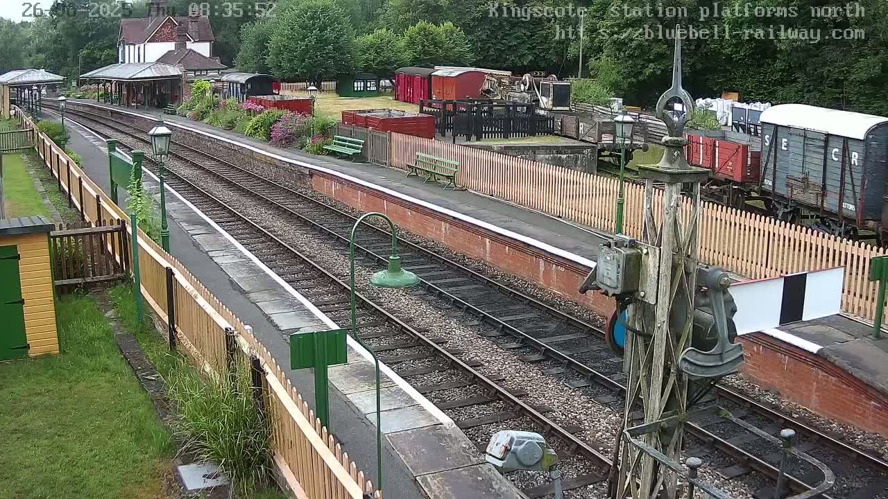 A train station platform with several railway cars and a station building visible, under overcast skies.