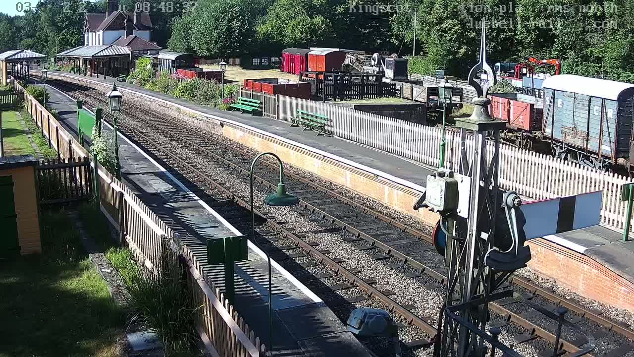 A sunny day at a train station with railway tracks, several freight cars, a station building, and wooden fences.