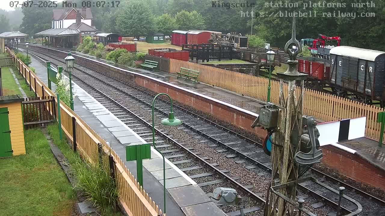 A wet railway station platform with multiple train tracks, a small station building, and several freight cars visible in the background.