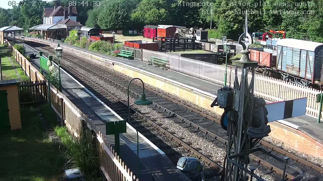 A sunny day at a railway station shows several train cars and a station building, with multiple train tracks extending into the distance.