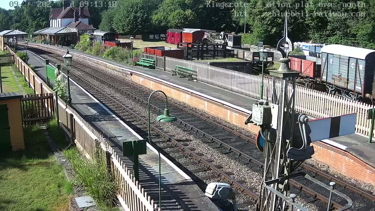 A sunny day at a train station shows multiple railway tracks, railway cars, and a station building surrounded by a wooden fence.