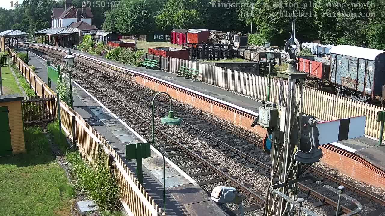 A train station platform with multiple train tracks, railway cars, and a station building under a sunny sky.
