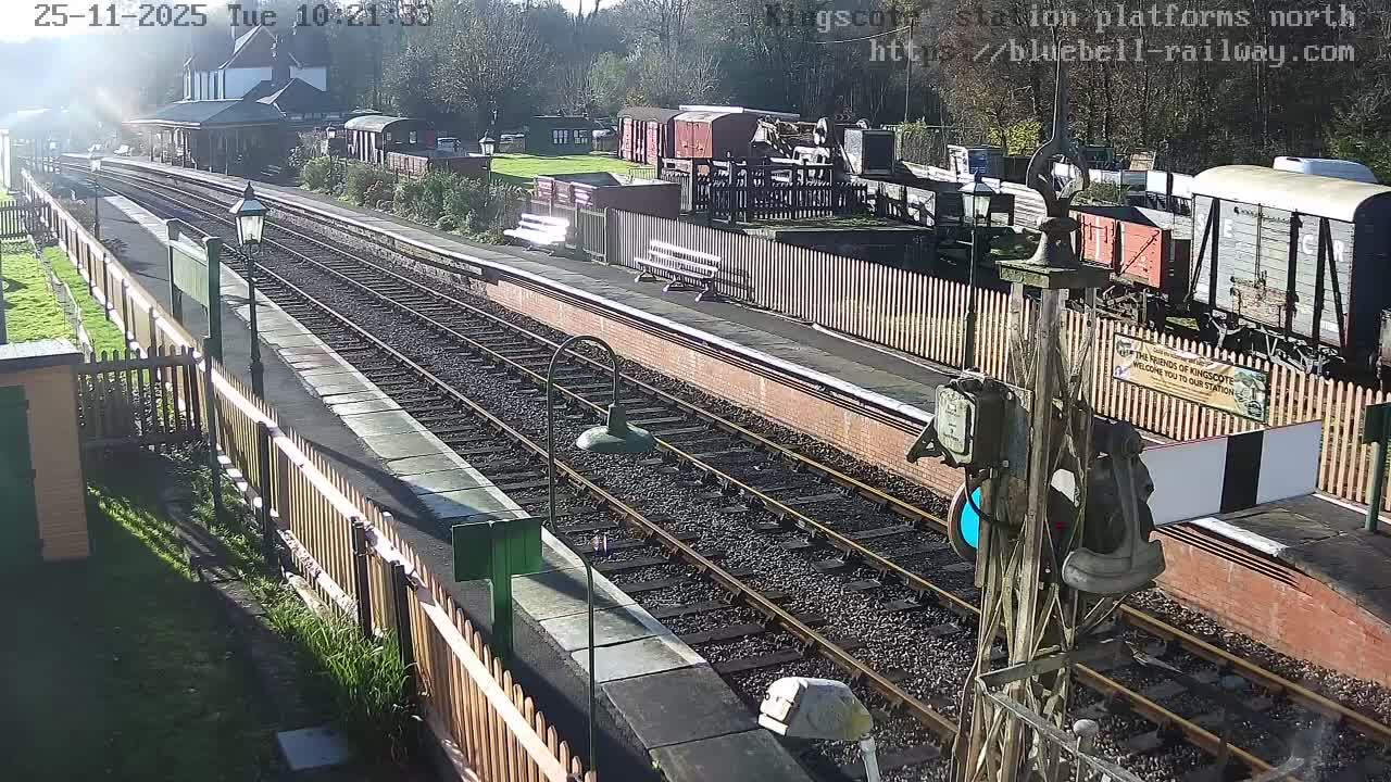 On a bright and sunny day, an empty, rural-style railway station features multiple tracks, platforms with vintage lampposts and benches, a station building, and several old train carriages parked on sidings.