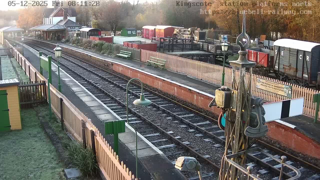 A picturesque, historic train station with multiple tracks, brick platforms, a main building, benches, and several parked railway wagons is seen on a bright, frosty morning under a clear sky.