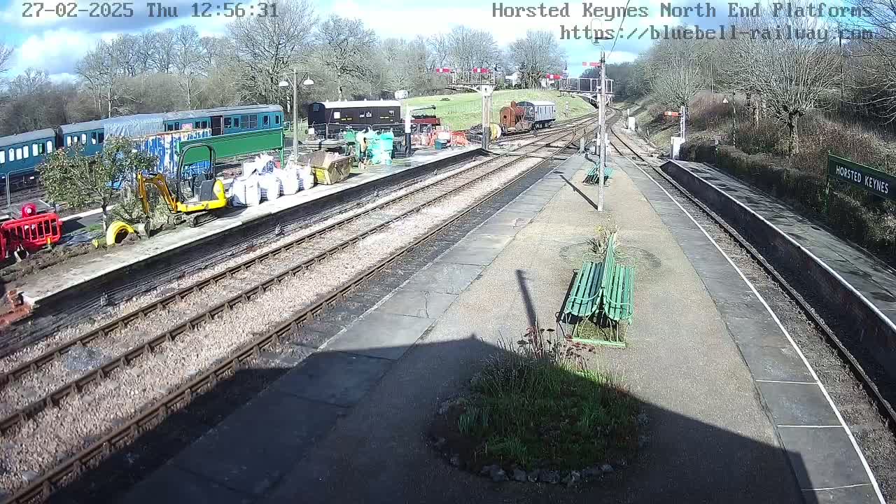 A sunny view of a train station platform with several train cars, construction equipment, and bags of material, alongside train tracks.