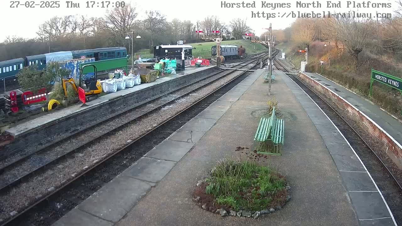 A train station with several train cars, construction equipment, and landscaping under an overcast sky.