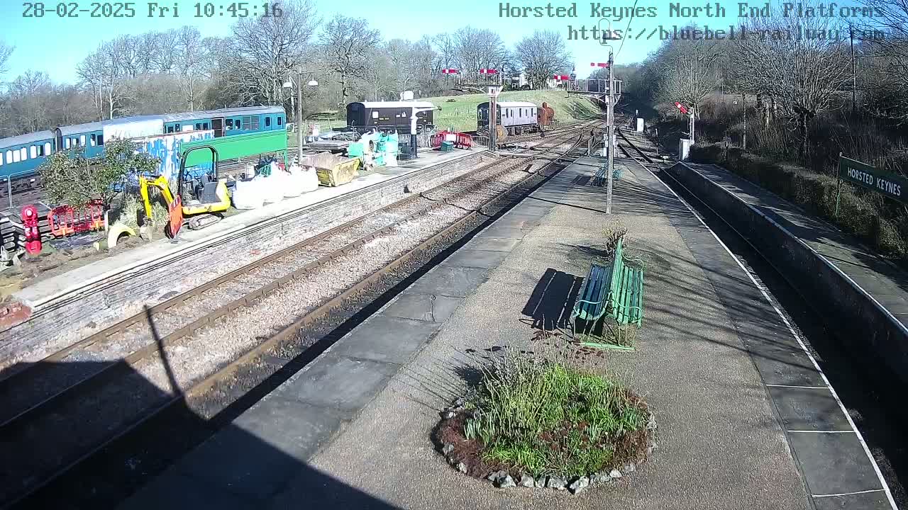 A sunny day at a train station with several railway cars parked on the tracks, construction equipment, and a small garden near benches.