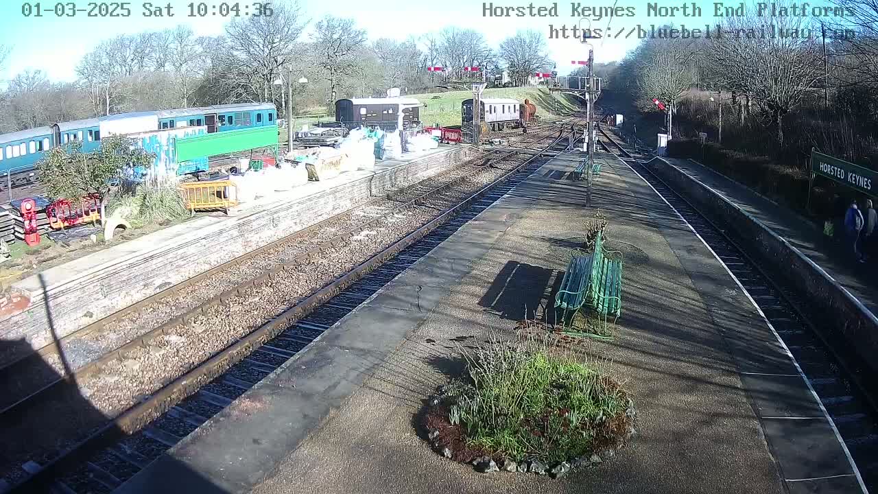 A sunny day at a train station shows several railway cars parked near a platform with benches and landscaping.