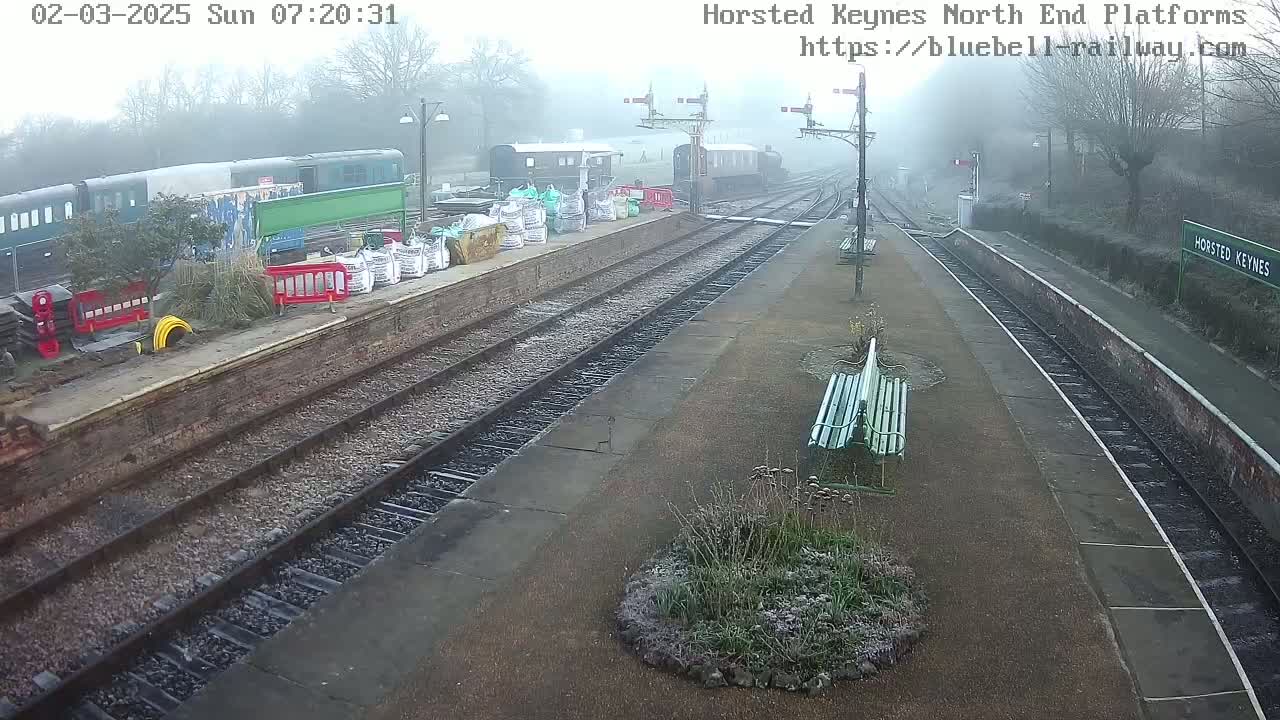 A foggy train station platform with several trains and construction materials visible, under a gray sky.