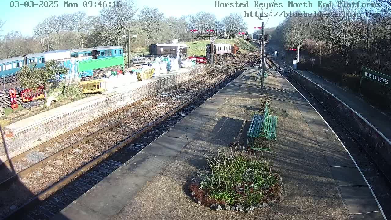 A sunny view of a train station platform with several railway carriages and construction materials visible, next to a grassy area with bushes and a bench.